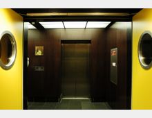 Innenraum eines Aufzugs mit dunklen Holzpaneelen an den Wänden, einer Metalltür in der Mitte und einer weißen beleuchteten Deckenverkleidung. Gelbe Aufzugstüren mit runden Fenstern sind teilweise geöffnet sichtbar.