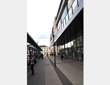 Straßenszene mit moderner Ladenpassage; links eine überdachte Wartezone mit Sitzbänken, rechts ein Gebäude mit großen Glasfassaden und Geschäften. Im Hintergrund sind mehrstöckige Wohnhäuser und ein gelbes Gebäude zu sehen.