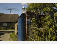 Eingang zu einem Privatgrundstück mit Ziegelmauer, blauem Tor und weißer Lampe an der Mauer. Im Hintergrund ist ein Gebäude mit Ziegeldach zu sehen, umgeben von Bäumen und Vegetation; darüber spannt sich eine Stromleitung.