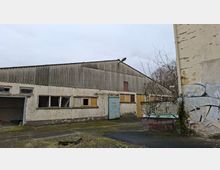 Verlassene Industriehalle mit Wellblechdach und verwitterter Fassade, teils zerbrochenen Fenstern und mehreren Türen an der Front. Davor liegt ein leerer, rissiger Hof mit Moosbewuchs, einer niedrigen Mauer mit Graffiti und kahlen Sträuchern am Rand.