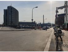 Straßenszene in Berlin mit Blick auf die Friedrichstraße; im Vordergrund eine Kreuzung mit Fahrrädern, Gehwegen und Straßenschildern. Im Hintergrund ein modernes Bürogebäude mit abgerundeten Ecken und der Bahnhof Friedrichstraße mit erkennbarem Gleisbereich.