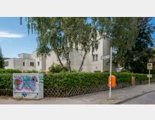 Straßenecke mit einem modernen Wohngebäude in Würfelform, umgeben von Bäumen und einer Hecke. Ein Stromkasten mit Graffiti, ein Straßenschild „Goldenes Horn“ sowie eine Bushaltestelle und ein orangefarbener Mülleimer sind sichtbar.