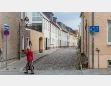 Gepflasterte Seitenstraße in einer Wohngegend mit hellen Reihenhäusern, teils geklinkert, und Satteldächern. Schilder für Verkehrsregeln und eine Laterne sind sichtbar; eine ältere Frau im roten Mantel überquert die Straße.