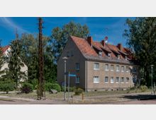 Zweistöckiges Mehrfamilienhaus mit hellbrauner Fassade und rotem Ziegeldach, umgeben von Bäumen und kleinen Grünflächen. Im Vordergrund Straßenlaternen, Straßenschilder und ein gepflasterter Gehweg.