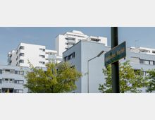 Mehrstöckige Wohngebäude in modernem Baustil mit hellblauen und weißen Fassaden im Märkischen Viertel. Im Vordergrund sind grüne Bäume und ein Straßenschild mit der Aufschrift „Märkisches Viertel“ an einem Laternenmast zu sehen.