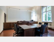 Ein Gerichtssaal mit dunklen Holzmöbeln, bestehend aus einem Richterpult, mehreren Schreibtischen und schwarzen Bürostühlen. An der rechten Seite befinden sich große Fenster, durch die Tageslicht einfällt, und links ein Holztürrahmen.