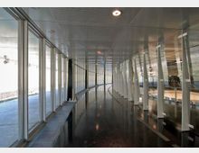 Ein moderner, lichtdurchfluteter Flur mit einer gebogenen Glasfassade, die von schmalen weißen Stützen unterteilt wird. Der dunkle, glänzende Boden spiegelt die Decke und die Umgebung wider, während die großen Fenster Tageslicht hereinlassen.