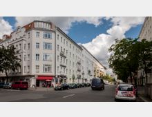 Straßenszene in einem Wohngebiet mit mehrstöckigen Altbauhäusern, die weiße Fassaden und vereinzelt Balkone haben. Am Straßenrand sind geparkte Autos, wenige Spaziergänger und ein kleines Straßencafé mit roten Markisen zu sehen, während Bäume den Gehweg säumen.