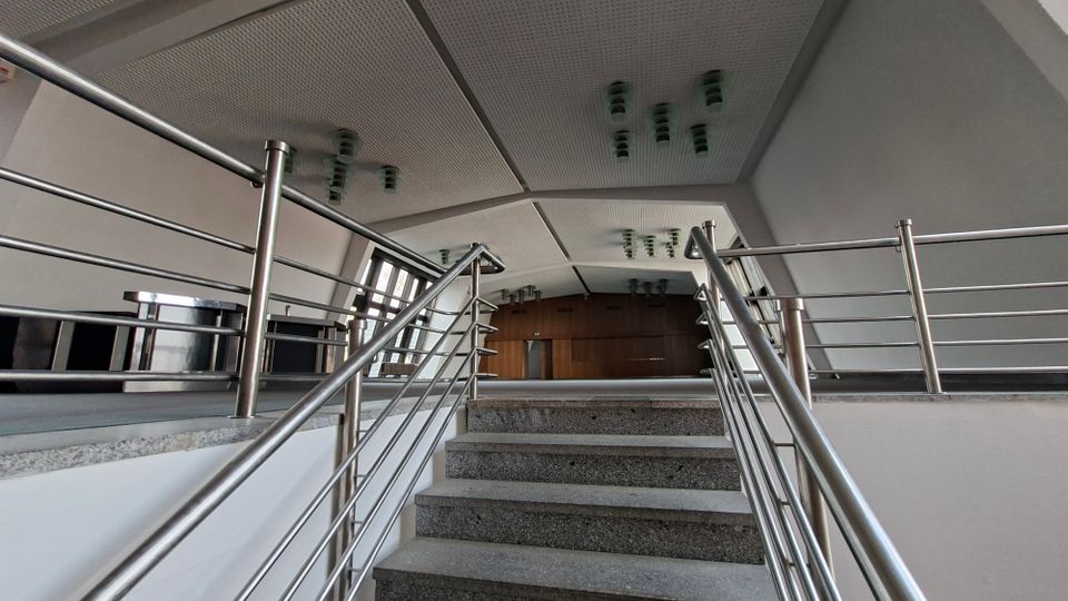 Innenansicht eines modernen Gebäudefoyers mit einer breiten Treppe aus grauem Stein und Edelstahlgeländern, die zu einer erhöhten Plattform führt. Darüber eine geneigte, paneelartige Decke mit mehreren Leuchten; links große Fenster und am Ende eine Holzverkleidung mit Türen.