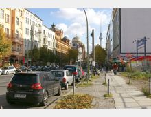 Straßenszene in Berlin mit parkenden Autos entlang einer mehrspurigen Straße. Auf der linken Seite stehen historische Gebäude, darunter die Neue Synagoge mit goldener Kuppel, während im Hintergrund der Berliner Fernsehturm sichtbar ist; rechts befinden sich moderne Gebäude und Gehwege mit Fußgängern.
