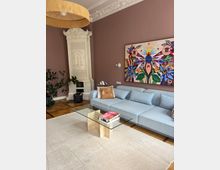 Elegantes Wohnzimmer mit hohen, mauvefarbenen Wänden, Stuckfries und einem dekorativen weißen Kachelofen in der Ecke. Vor einem großen, farbenfrohen Wandbild stehen ein hellblaues Sofa, ein gläserner Couchtisch auf hellem Teppich sowie eine große Hängeleuchte und Parkettboden.
