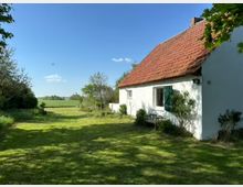 Weißes Landhaus mit rotem Ziegeldach und grünen Fensterläden, umgeben von einem weitläufigen Garten mit Rasen und Bäumen, im Hintergrund offenes Feld unter klarem, blauem Himmel.