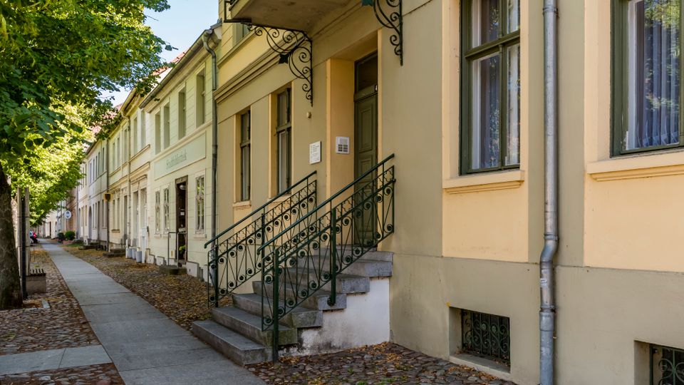 Straßenzug mit alten, hell gestrichenen Wohnhäusern und hohen Fenstern, davor ein Gehweg entlang einer Baumallee. Im Vordergrund führt eine Treppe mit schmiedeeisernem Geländer zu einem Hauseingang; Kopfsteinpflaster und fallendes Laub säumen den Rand.