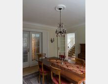 Esszimmer mit einem Holztisch, vier gepolsterten Stühlen und einem rot-goldenen Tischläufer. Im Raum befinden sich ein Kronleuchter, eine Holzkommode und Türen mit Glasflächen; im Hintergrund ist eine Treppe sichtbar.