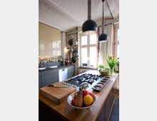 Moderne Küche mit einer Kücheninsel aus Holz, die ein Gasherd und Gewürzbehälter integriert. Im Hintergrund befinden sich graue Schränke, eine Spüle, hängende Töpfe und Pfannen an einer Ziegelwand sowie große Fenster, die den Raum mit Tageslicht erhellen.