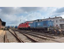 Bahnhofsgelände mit mehreren Gleisen und geparkten Lokomotiven, darunter eine rote und eine blaue. Im Hintergrund sind ein Backsteingebäude, moderne Gebäudefassaden und ein wolkiger Himmel zu sehen.