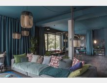 Ein modern eingerichteter Wohnraum mit einer großen grauen Couch, die mit bunten Kissen dekoriert ist, sowie mehreren hängenden Lampen aus Holz. Der Raum ist von dunkelblauen Vorhängen geprägt, hat große Fenster mit Blick ins Grüne und eine gemütliche Atmosphäre mit Pflanzen und dekorativen Elementen.