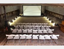 Ein kleiner Veranstaltungsraum mit einer weißen Projektionsleinwand an der Stirnseite, umgeben von rauen, metallischen Wänden. Mehrere Reihen weißer Klappstühle stehen auf einem Holzboden, flankiert von dezentem Bodenlicht entlang der Seitenwände.