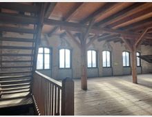 Weitläufiger Innenraum eines historischen Gebäudes mit sichtbaren Holzbalken, Holztreppe und einer Reihe hoher Fenster mit schlichtem Metallrahmen. Die Wände bestehen aus alter, teilweise verputzter Ziegelstruktur, und der Boden ist aus Holz.