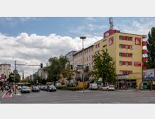 Straßenszene mit mehrstöckigen Wohn- und Geschäftshäusern, darunter ein gelbes Gebäude mit roten Akzenten und einem Drogeriemarkt im Erdgeschoss. Im Vordergrund sind Autos an einer Kreuzung mit Verkehrsschildern, Bäumen und Gehwegen zu sehen.