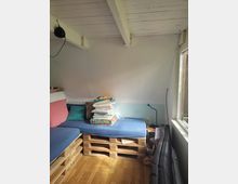 Kleines Zimmer mit schräger weißer Holzdecke und sichtbaren Balken, zwei niedrigen Betten aus Europaletten mit blauen Matratzen und mehreren Kissen. Rechts fällt Tageslicht durch ein großes Fenster; daneben stehen eine kleine Holz-Nachttischablage, eine Leselampe und ein gepolstertes Möbel an der Wand.