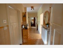 Schmaler Wohnungsflur mit hellem Fischgrät-Parkett, beige gestrichenen Wänden und mehreren weißen Türen entlang des Gangs. An der Wand stehen eine Holzkommode und ein Wandspiegel über einem kleinen Regal neben einem Heizkörper; am Ende führt eine geöffnete Tür in einen weiteren Raum.