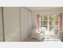 Helles Wohnzimmer mit bodentiefen weißen Fenstern, die von rosa Vorhängen eingerahmt sind, einer weißen Sesselgruppe und einem gläsernen Beistelltisch. Rechts ist eine Glas-Schiebetür zu sehen, die einen Einbauschrank verdeckt.