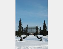 Ein großer, schneebedeckter Gartenweg führt zu einem gläsernen Gewächshaus mit einer Kuppelstruktur, flankiert von hohen, symmetrisch angeordneten Nadelbäumen und dekorativen Pflanzgefäßen. Der Himmel ist klar und blau, was die winterliche Szene unterstreicht.