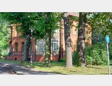 Backsteingebäude im Stil der Gründerzeit, umgeben von hohen Bäumen und Grünflächen. Ein Straßenlaterne und ein Verkehrsschild mit Parkhinweis sind im Vordergrund sichtbar.