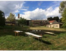 Offene Grünfläche mit vereinzelten Tischen und Stühlen, im Hintergrund mehrere alte Gebäude aus Stein und Ziegel mit teils wellblechgedeckten Dächern. Bäume und bewölkter Himmel ergänzen die ländliche Szenerie.