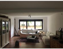 Wohnzimmer mit schlichtem Mobiliar, darunter zwei weiße Sofas, ein kleiner Holztisch und ein antiker Holzschrank links. Große Fenster im Hintergrund bieten Blick auf eine Gartenlandschaft, während ein dunkler Teppich den Raum akzentuiert.