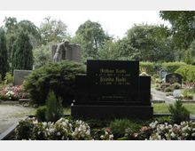 Gräberfeld auf einem Friedhof mit gepflegten Blumenbeeten, einem dunklen Grabstein mit goldener Inschrift und einer steinernen Trauerstatue im Hintergrund. Umgeben von Bäumen und Sträuchern in einer ruhigen, grünen Umgebung.
