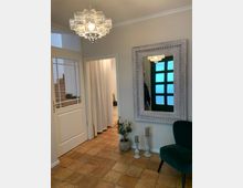 Eingangsbereich mit beigem Fliesenboden, einer weißen Tür mit Glaselementen und einer Wand mit einem großen silbergerahmten Spiegel. Ein auffälliger, orientalisch verzierter Kronleuchter hängt von der Decke, daneben befinden sich grüne Dekorationen und Kerzenhalter auf dem Boden.