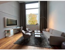 Wohnraum mit modernem Design: Ein graues Sofa, zwei Armstühle und ein kleiner Couchtisch stehen auf einem Teppich mit Holzfußboden. Eine große Fensterfront mit Vorhängen lässt Tageslicht herein, daneben ein Schreibtisch mit Fernseher an der Wand.