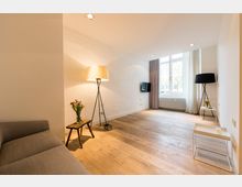 Modernes Wohnzimmer mit Holzboden und weiß gestrichenen Wänden, ausgestattet mit einer grauen Couch, Stehlampen, einem kleinen Tisch mit Blumenstrauß und einem an der Wand montierten Flachbildfernseher. Große Fenster mit Vorhängen lassen Tageslicht herein, daneben befindet sich ein Heizkörper.