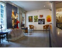 Ein kleiner Galerieraum mit hellen Wänden, auf denen mehrere farbenfrohe Gemälde hängen, darunter Darstellungen eines Schwans, einer Flamme und Naturmotive. Links befindet sich eine Glasvitrine mit einer Puppenausstellung, während im Raum Rattanstühle und kleine Tische stehen.