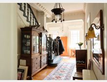 Ein traditionelles, helles Interieur mit einer Holztreppe, verziertem Geländer und geprägter Deckenleiste. Links ein antiker Holzschrank mit Glastüren, rechts ein Wandspiegel und eine Tischlampe; in der Mitte führt ein bunter Teppich durch den Raum.