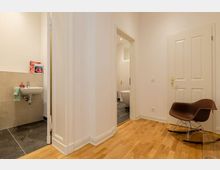 Heller Wohnungsflur mit Holzboden, einer braunen Schaukelstuhl-ähnlichen Sitzgelegenheit und weiß gestrichenen Wänden. Im Hintergrund befinden sich ein Durchgang zu einem Badezimmer mit freistehender Badewanne sowie eine separate Nische mit Waschbecken und dunklen Fliesen.