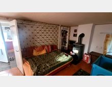 Kleines Wohnzimmer mit gemusterten Tapeten, einem Sofa mit Decken und Kissen, einem Holzofen, einem Bücherregal und einer Tür zu einem angrenzenden Raum. Der Boden ist mit roten Fliesen bedeckt, und an der rechten Seite befindet sich ein Vorhang sowie eine Spielbox.