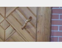 Großaufnahme einer hölzernen Tür mit diagonalem Muster und einem geschwungenen Türgriff aus Holz. Im Hintergrund ist teilweise eine Wand mit rotem Ziegelmuster zu sehen.