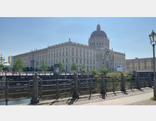 Historisches Gebäude mit einer großen Kuppel, bekannt als das Humboldt Forum, im Stil eines Barockschlosses. Im Vordergrund fließt ein Kanal mit steinernem Geländer, umgeben von Bäumen und modernem Stadtmobiliar.