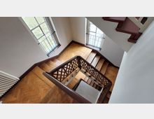 Treppenhaus mit einer hölzernen Treppe und verzierten Geländern, das durch zwei große Fenster mit Gitterstäben beleuchtet wird. Der Boden ist aus Holz im Fischgrätmuster verlegt, und die Wände sind schlicht gehalten.