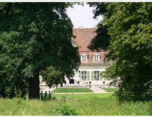 Historisches Haus mit rotem Ziegeldach und grünen Fensterläden, eingebettet in eine parkähnliche Landschaft mit großen Bäumen und Rasenflächen. Im Vordergrund stehen Menschen unter einem Baum, während der Eingangsbereich des Gebäudes im Hintergrund sichtbar ist.