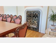 Ein eleganter Innenraum mit einem großen Holztisch und gepolsterten, ornamentverzierten Stühlen. Im Hintergrund ist ein Kamin mit einer dekorativen goldenen Gittertür zu sehen, daneben steht eine grüne Topfpflanze.