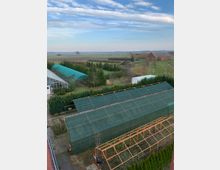 Blick auf eine ländliche Landschaft mit Feldern, Bäumen und vereinzelten Gebäuden im Hintergrund. Im Vordergrund sind mehrere Gewächshäuser und eine Struktur mit Metallgestell zu sehen.