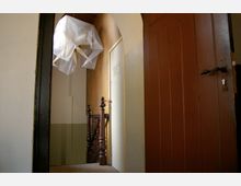 Ein schmaler Innenflur mit einem hölzernen Treppengeländer und einer geschlossenen weißen Tür. Über der Treppe hängt eine mit Plastikfolie abgedeckte Lampe, und rechts ist eine geöffnete braune Holztür sichtbar.