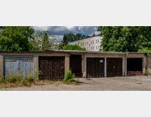 Reihe alter Garagen mit Holz- und Metalltoren, umgeben von vereinzeltem Bewuchs und Bäumen im Hintergrund. Im oberen Bereich sind Wohnhäuser mit hellen Fassaden sichtbar.