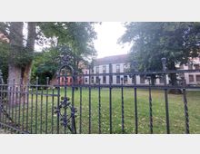 Eiserner Zaun mit detaillierten Ornamenten, der eine Grünfläche mit großen Bäumen umgibt, dahinter ein zweistöckiges Gebäude mit roten und weißen Fassaden sowie einer Ziegeldachbedeckung.