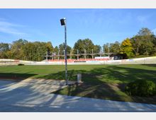 Ein Freiluft-Radstadion mit einer Betonbahn und einem zentralen Rasenbereich, umgeben von Tribünen mit roten Sitzen und einem überdachten Bereich. Im Hintergrund befinden sich Bäume und Werbebanner entlang der Bahnbegrenzung.