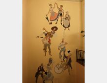 Wandgemälde in einer Treppenhausnische mit Figuren in traditioneller und historischer Kleidung, darunter Tänzer, ein Musiker mit Gitarre, eine Frau mit Regenschirm und ein Jongleur mit Reifen. Die Szene ist auf einer hellgelben Wand dargestellt, begleitet von einem schmiedeeisernen Treppengeländer rechts.
